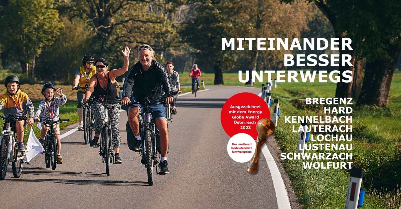 Mehrere Erwachsene und Kinder fahren fröhlich mit dem Fahrrad auf einer Landstraße, winken in die Kamera und wirken ausgelassen. Rechts im Bild steht der Schriftzug ‚Miteinander besser unterwegs‘ sowie die Namen der acht plan b-Gemeinden. Zusätzlich ist ein rotes Siegel mit dem Hinweis ‚Ausgezeichnet mit dem Energy Globe Award Österreich 2023‘ zu sehen.