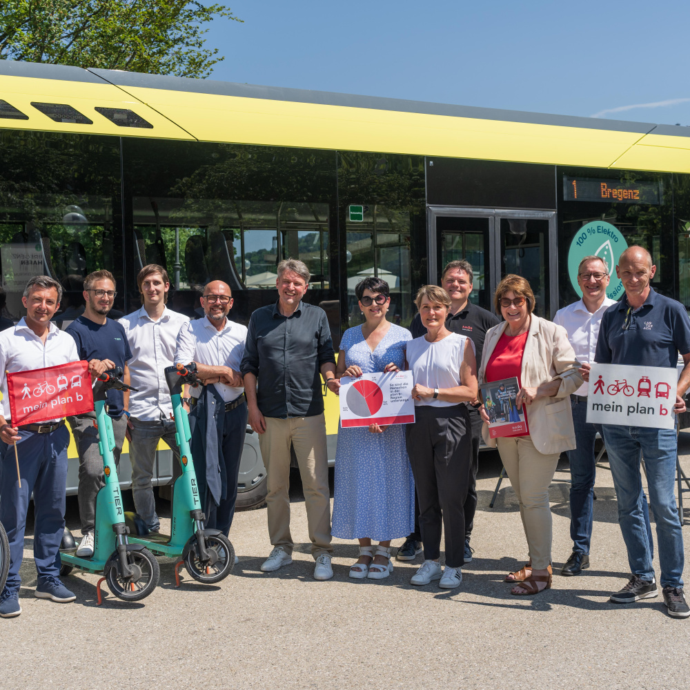 Vertreter:innen der plan b-Gemeinden stehen mit E-Scootern, Fahrrädern und Plakaten zu plan b vor einem E-Bus des Landbus Unterland bei schönem Wetter beim Platz der Menschenrechte in Bregenz