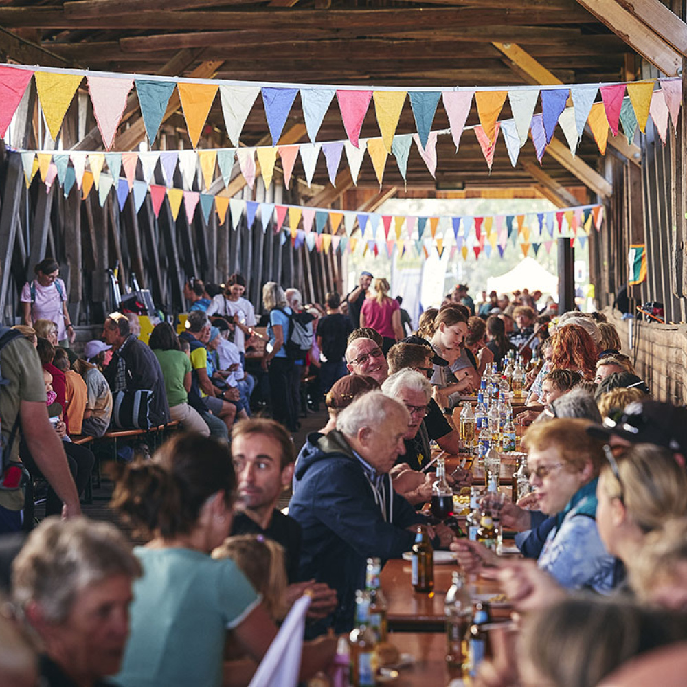 Menschen genießen in der alten Senderbrücke kulinarische Genüsse beim plan b-Rad-Ried-Tag. Die alte Senderbrücke ist geschmückt mit bunten Fahnen die von der Decke hängen.