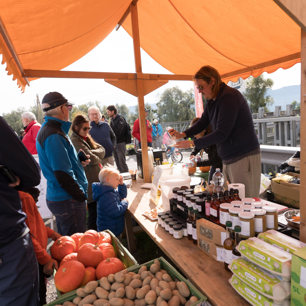 Besucher:innen kaufen bei einem Bauernmarkt Köstlichkeiten aus der Region wie Kartoffeln, Kürbisse oder Honig.