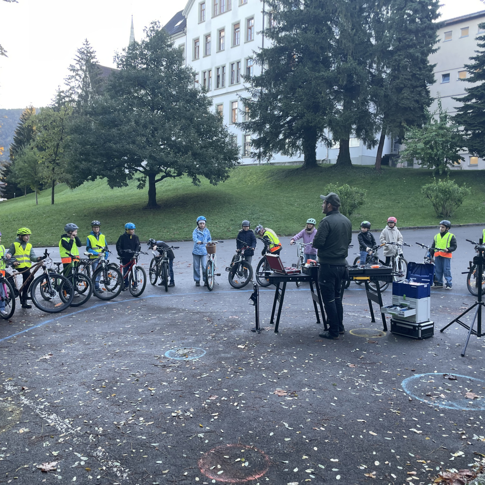 Kinder einer Volksschulklasse stehen beim Schulplatz rund um den Rad-Mechaniker der mit ihnen einen Rad-Reparatur-Kurs durchführt