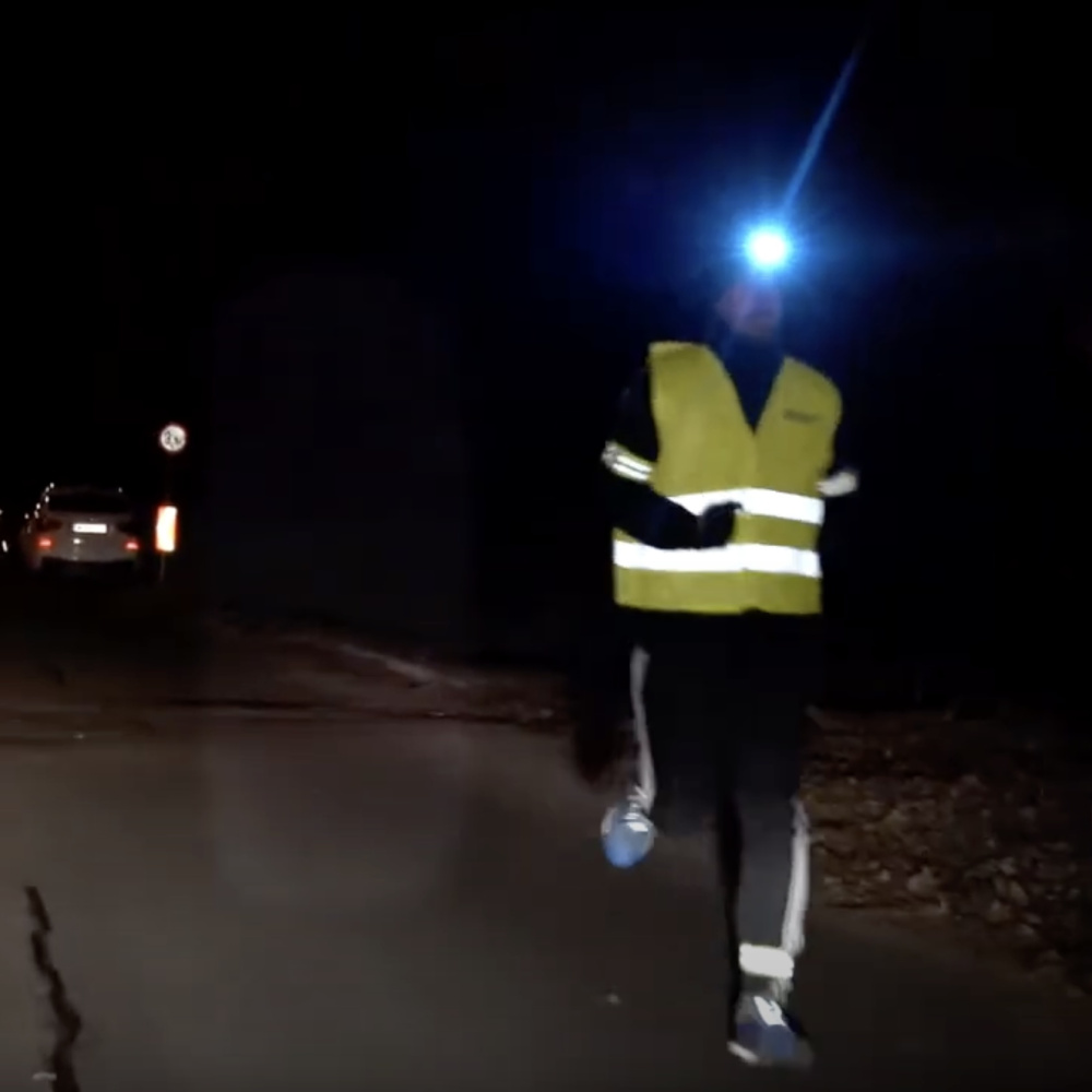 Eine Person joggt in der Dunkelheit und macht sich mit Warnweste, Stirnlampe und Refelektorbändern sichtbar.