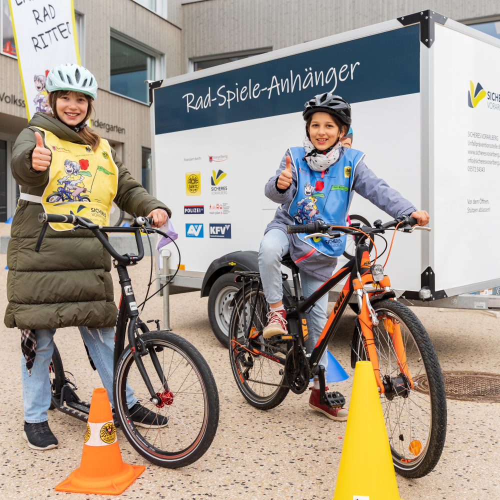 Zwei Volksschüler:innen stehen mit Ihrem Fahrrad vor dem Rad-Spiele-Anhänger und zeigen Ihre Freude per "Daumen hoch"