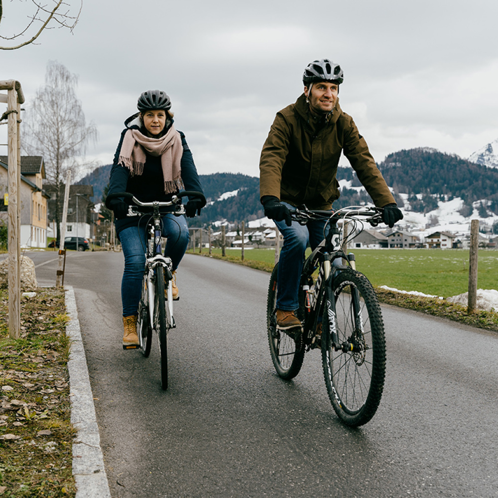 Zwei Personen sind im Winter per Rad unterwegs. Sie sind warm angezogen. Im Hintergrund sind schneebedeckte Berge zu sehen.