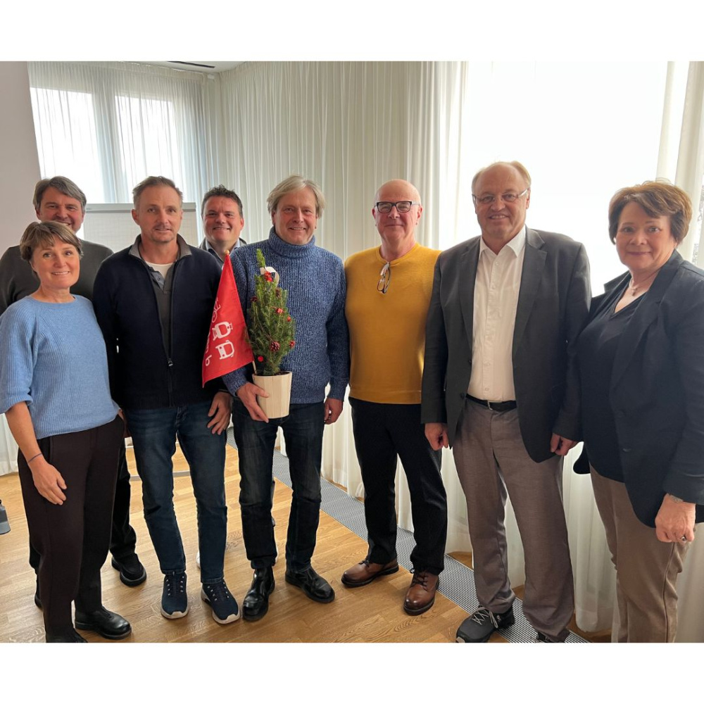 Gruppenfoto mit den Vertreter:innen der plan b-Gemeinden im Sitzungssaal der Gemeinde Lauterach. In der Mitte der Bürgermeister Frank Matt mit Geschenk zum Beitritt zu den plan b-Gemeinden. 