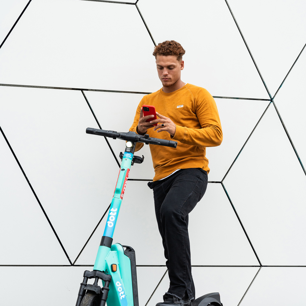 Ein Mann mit organgem mPulli  steht mit seinem Handy vor einem türkisen E-Scooter und möchte ihn buchen