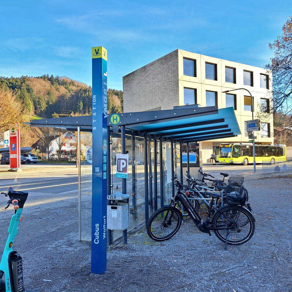 VMOBIL Station Wolfurt Cubus mit E-Scooter, Fahrradabstellplatz und Bus im Hintergrund
