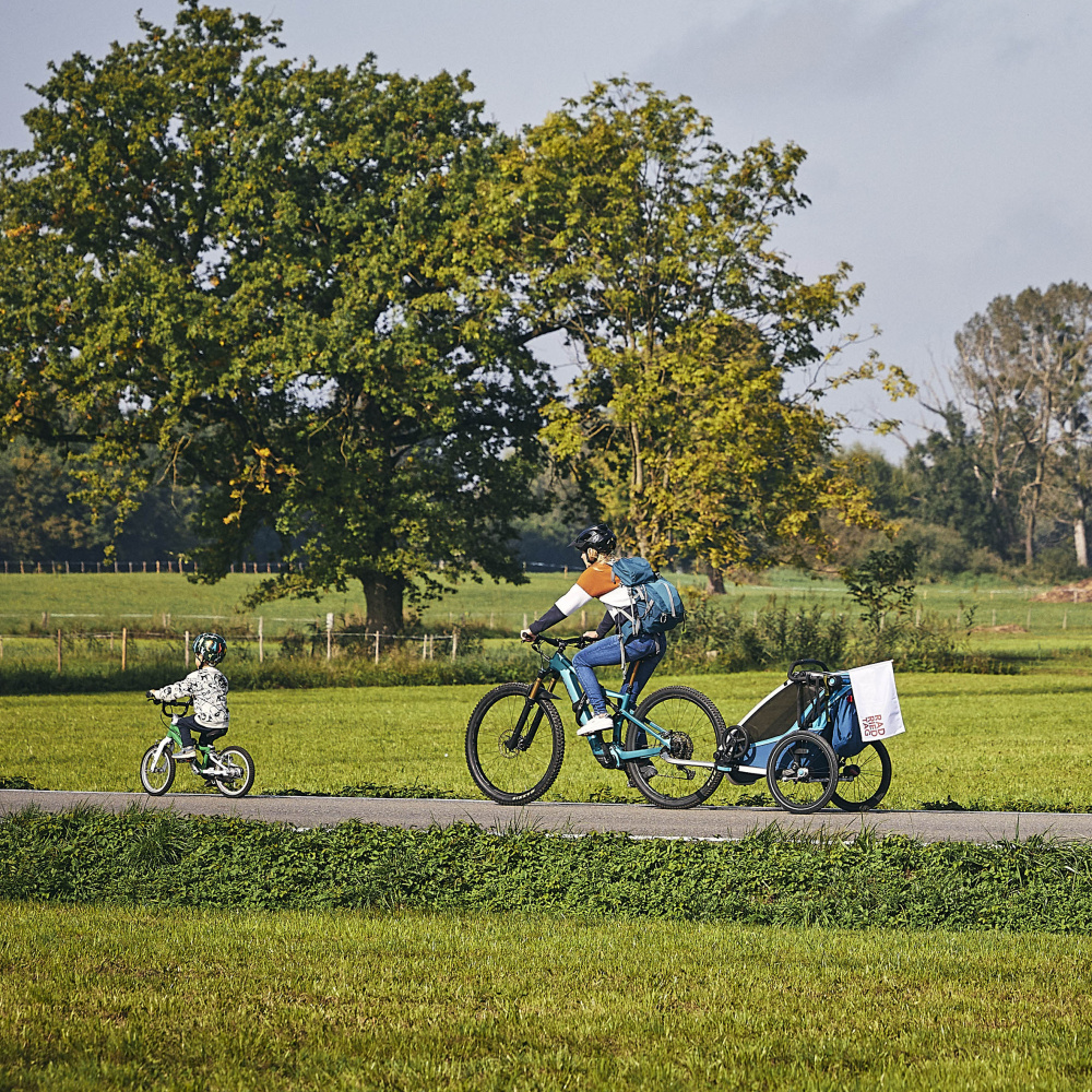 Eine Frau mit Fahrrad und Kiki und ein kleiner Junge mit Fahrrad fahren gemeinsam auf den autofreien Straßen im Ried bei schönem Wetter und grüßen Wiesen und Bäumen
