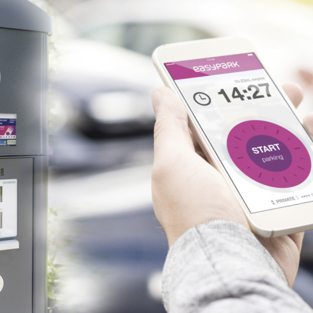 Hand mit Handy vor einem Parkautomaten. Es wird die App "EasyPark" angezeigt