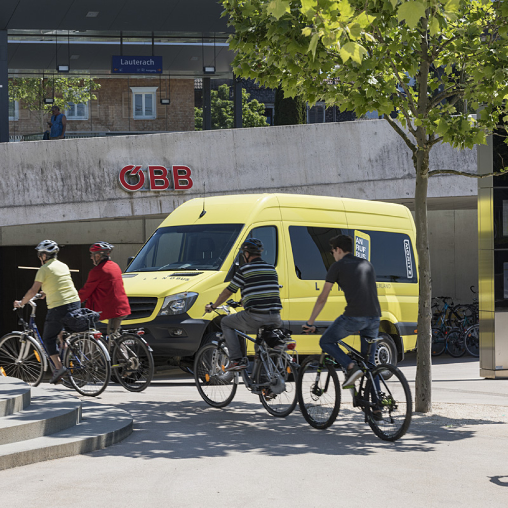 Mehrere Radfahrer:innen fahren beim Bahnhof in Lauterach vor dem gelben Anrufbus vorbei. Im Bild sind Bäume sowie eine Treppe zu sehen.