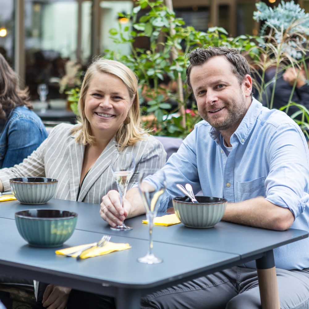 Zwei Personen sitzen lächelnd an einem gedeckten Tisch im Freien und genießen ein Essen