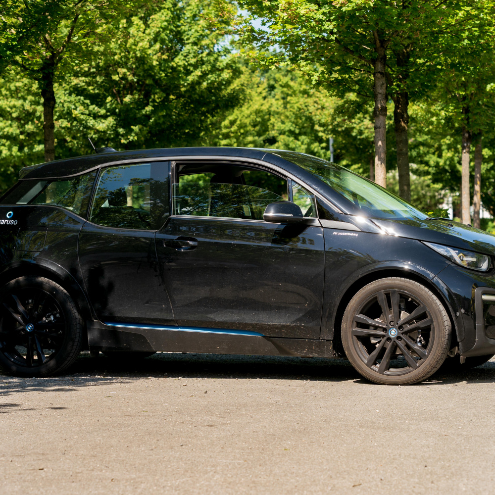 Schwarzes E-Auto an einer Ladestation. Im Hintergrund sind grüne Bäume zu sehen. 