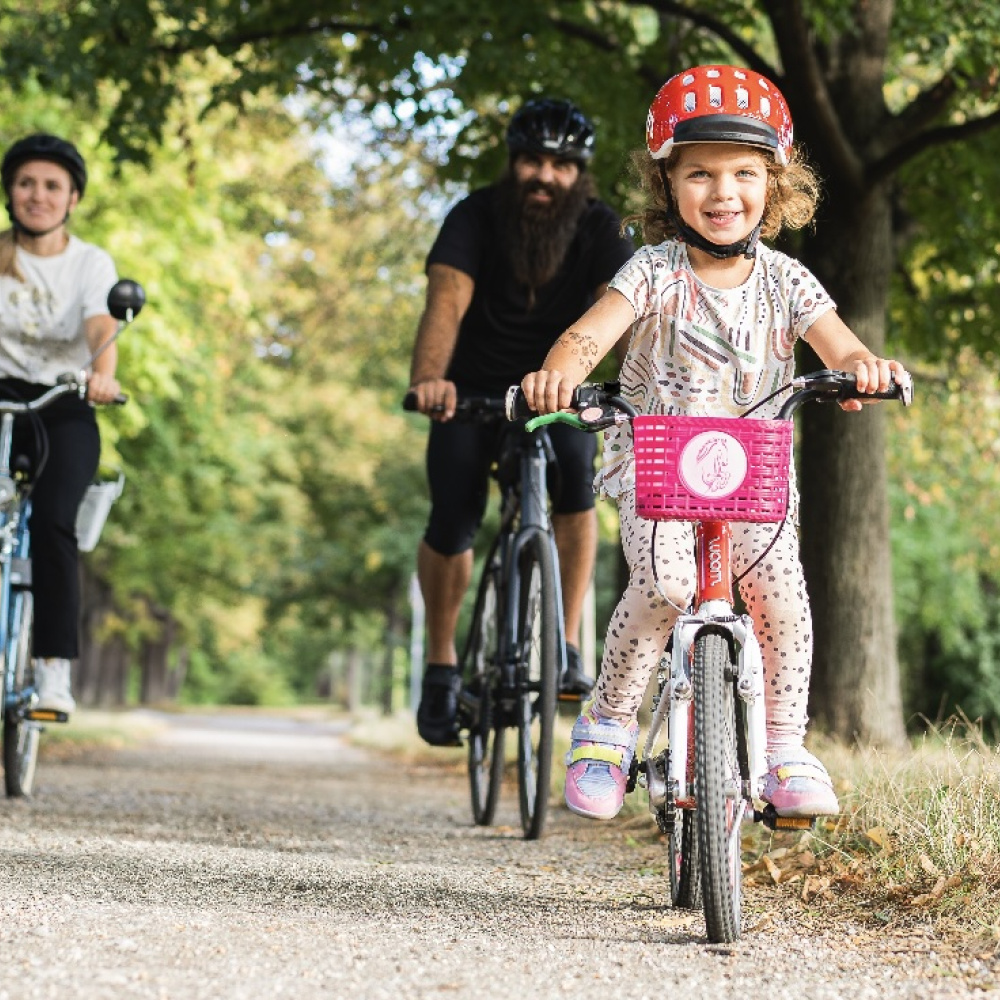 Ein fröhliches Mädchen mit rotem Helm fährt auf einem kleinen Fahrrad mit pinkem Korb an einem sonnigen Tag in einer grünen, baumbestandenen Allee ab