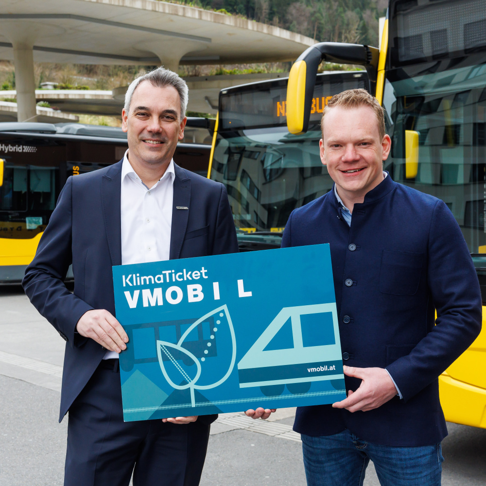 VVV Geschäftsführer Christian Hillbrand und Landestatthalter Christof Bitschi halten ein großes Klimaticket vor Bussen am Bahnhof.