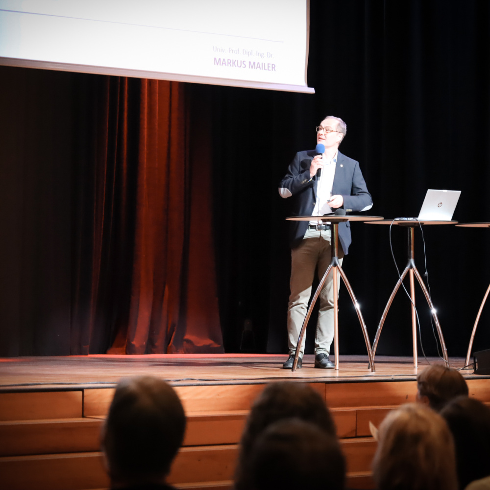 Professor Markus Mailer präsentiert auf der Bühne per Präsentation. Im Vordergrund sind die Zuschauer:innen zu sehen. 