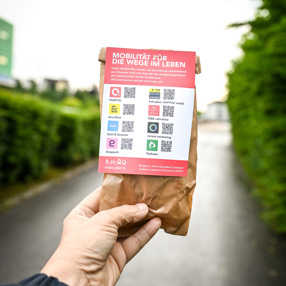 Eine Hand hält eine braune Papiertüte mit einem roten Infokärtchen, das verschiedene Mobilitätsangebote und QR-Codes zeigt. Im Hintergrund verläuft ein asphaltierter Weg zwischen grünen Hecken. In der Tüte befindet sich ein Zopf