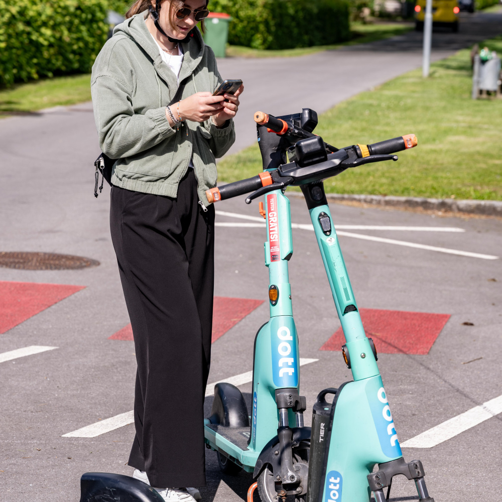 Eine Frau mit Helm und Sonnenbrille steht mit Ihrem Handy vor zwei türkisen Leih-E-Scootern und möchte einen der Fahrzeuge ausleihen.