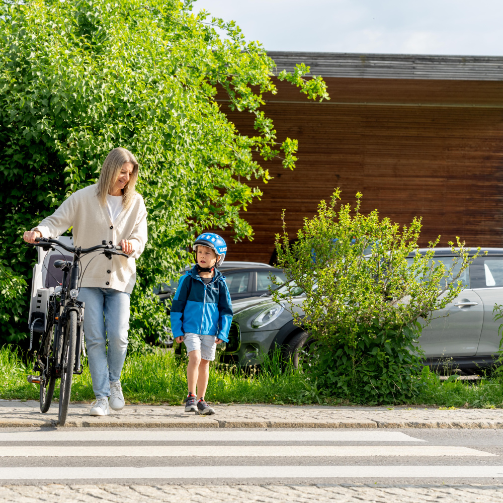 Eine Frau schiebt ihr Fahrrad über einen Zebrastreifen und begleitet dabei einen kleinen Jungen mit Fahrradhelm. Beide stehen am Straßenrand und wollen gemeinsam die Straße überqueren. Im Hintergrund sind grüne Sträucher, ein silbernes Auto und ein Holzgebäude zu sehen. Ein Verkehrsschild markiert die Fußgängerüberquerung