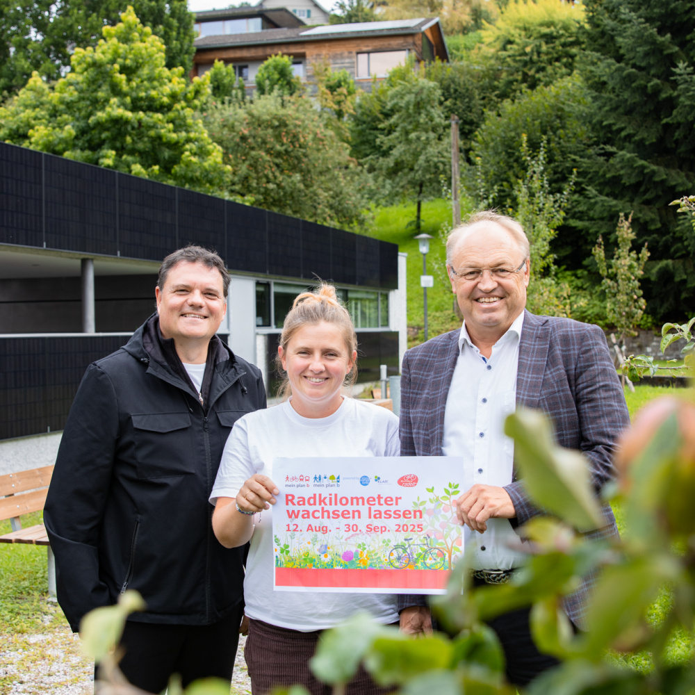 Bgm. Thomas Schierle, Julie Buschbaum und Bgm. Elmar Rhomberg stehen im Grünen und laden gemeinsam mit dem Plakat "Radkilometer wachsen lassen von 12. August - 30. September" zur Aktion. Im Vordergrund sind unscharf einige Äpfel am Baum zu sehen. Die Stimmung ist freundlich und naturverbunden.