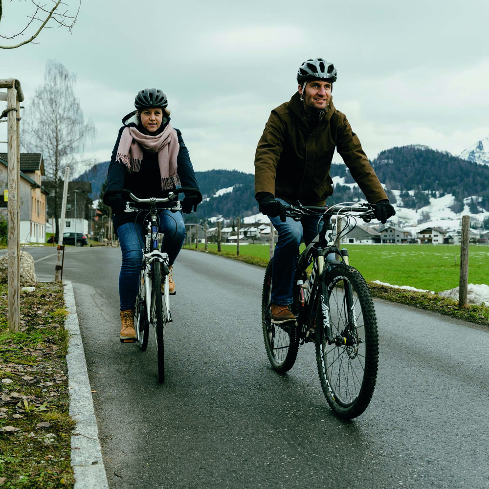 Zwei Personen sind warm gekleidet im Winter mit dem Fahrrad unterwegs. Im Hintergrund verschneite Berge. Die Straßen sind frei von Schnee. 