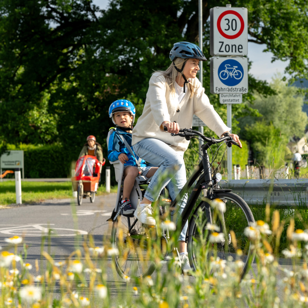 Frau fährt mit Kind auf dem Fahrrad entlang eines Radwegs. Im Vordergrund eine Blumenwiese.