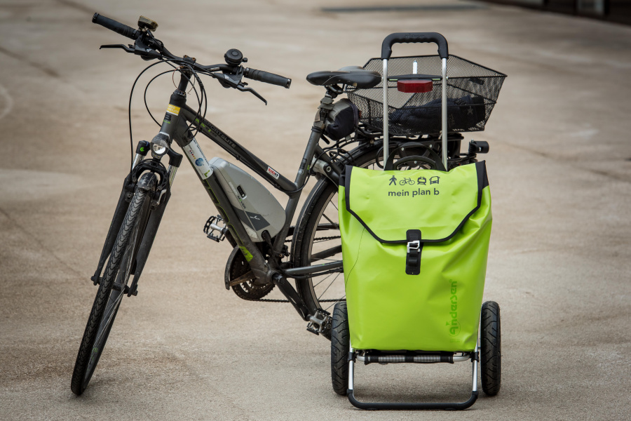 Ein schwarzes Fahrrad auf Asphalt, daneben steht ein grüner Fahrrad-Trolley mit plan b-Logo