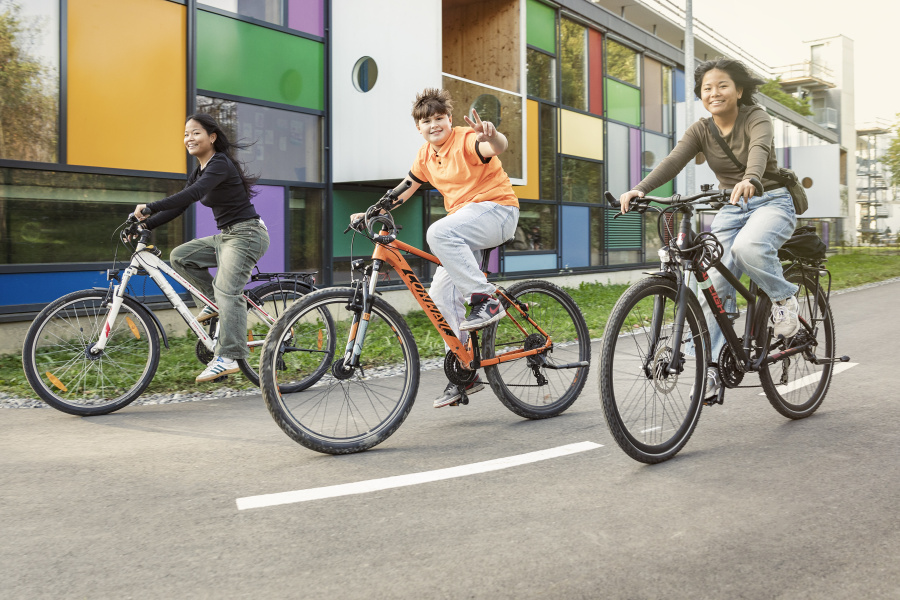 Drei gut gelaunte Jugendliche unterwegs auf dem Fahrrad vor einem bunten Gebäude 