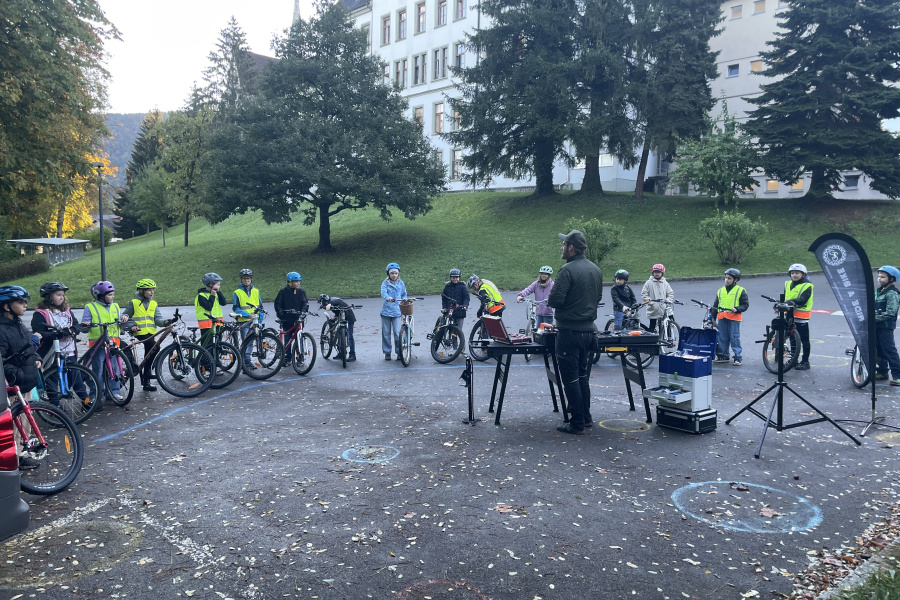 Kinder einer Volksschulklasse stehen beim Schulplatz rund um den Rad-Mechaniker der mit ihnen einen Rad-Reparatur-Kurs durchführt