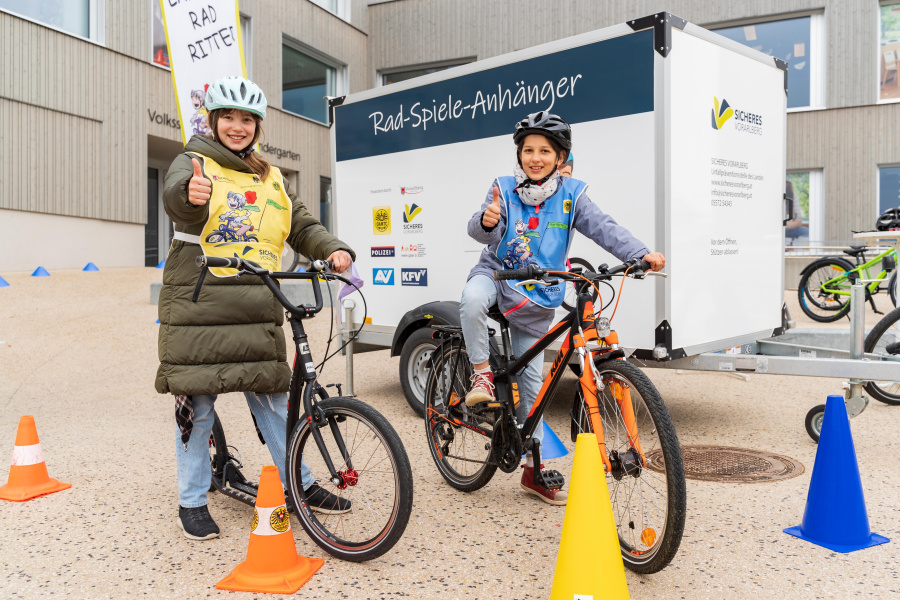 Zwei Volksschüler:innen stehen mit Ihrem Fahrrad vor dem Rad-Spiele-Anhänger und zeigen Ihre Freude per "Daumen hoch"