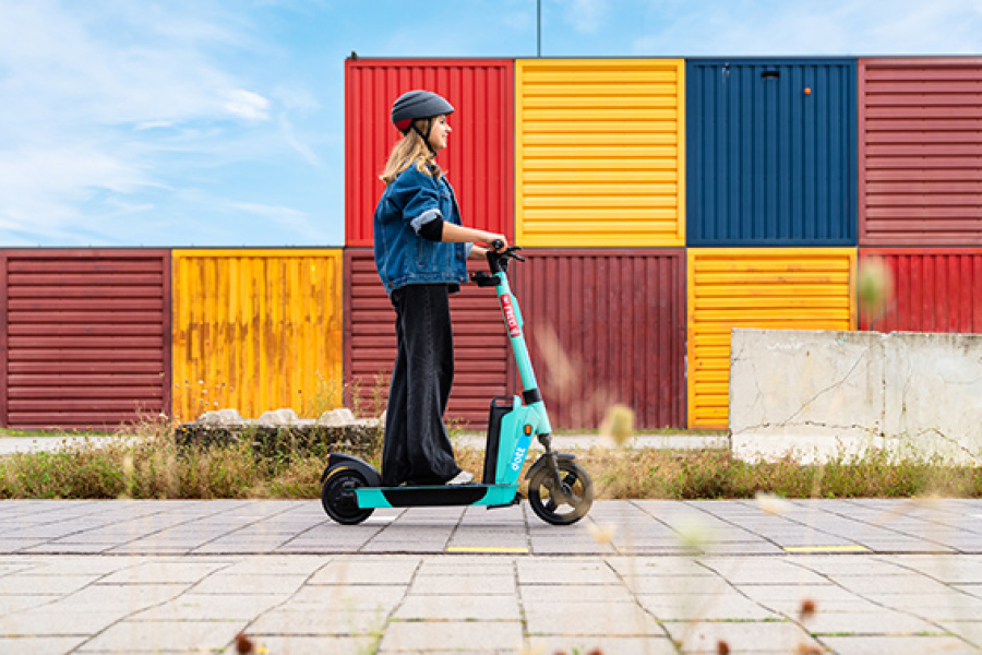 Frau mit Helm fährt mit dem E-Scooter auf einer Straße. Im Hintergrund stehen bunte Container. 