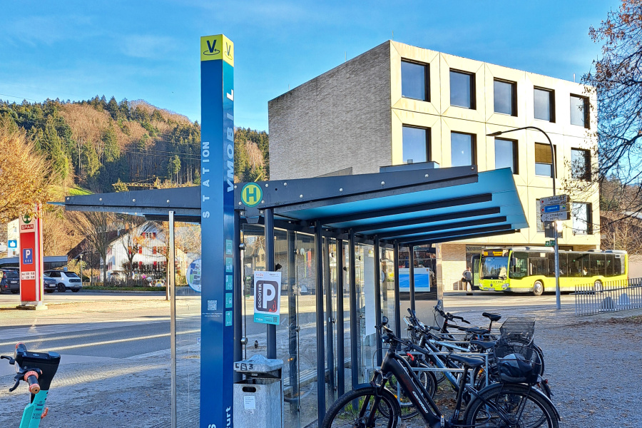 VMOBIL Station Wolfurt Cubus mit E-Scooter, Fahrradabstellplatz und Bus im Hintergrund