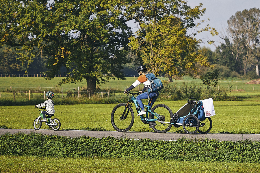 Eine Frau mit Fahrrad und Kiki und ein kleiner Junge mit Fahrrad fahren gemeinsam auf den autofreien Straßen im Ried bei schönem Wetter und grüßen Wiesen und Bäumen