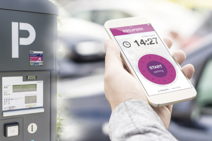 Hand mit Handy vor einem Parkautomaten. Es wird die App "EasyPark" angezeigt
