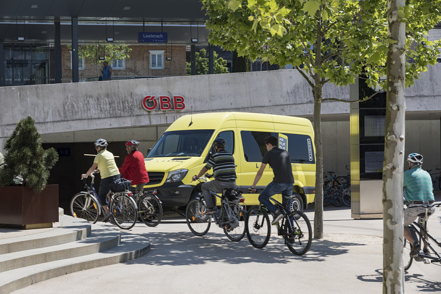Mehrere Radfahrer:innen fahren beim Bahnhof in Lauterach vor dem gelben Anrufbus vorbei. Im Bild sind Bäume sowie eine Treppe zu sehen. 