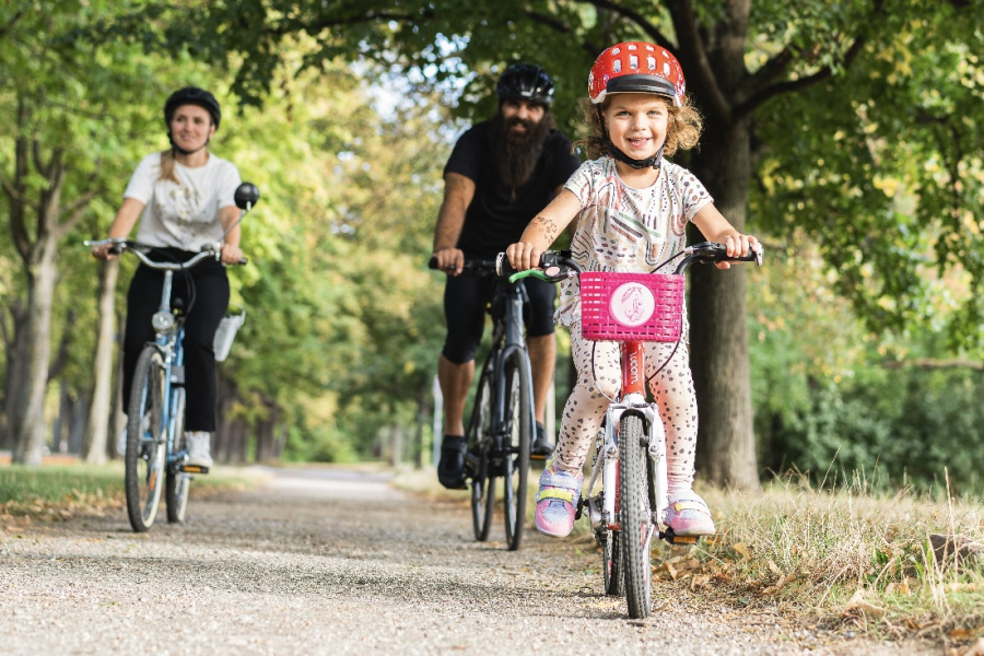 Ein fröhliches Mädchen mit rotem Helm fährt auf einem kleinen Fahrrad mit pinkem Korb an einem sonnigen Tag in einer grünen, baumbestandenen Allee ab