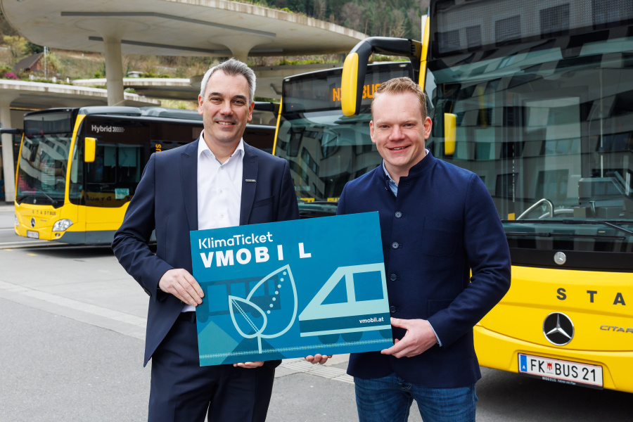 VVV Geschäftsführer Christian Hillbrand und Landestatthalter Christof Bitschi halten ein großes Klimaticket vor Bussen am Bahnhof.