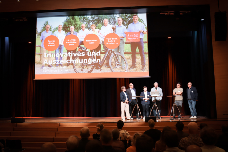 Die Vertreter:innen stehen auf der Bühne im Saal. Dort läuft die Präsentationsfolie. Unten auf den Stühlen sitzen die Zuschauer:innen. 