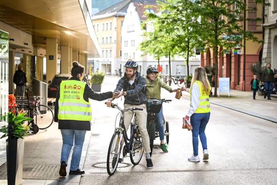 Zwei Radfahrer:innen erhalten von den plan b-Vertreterinnen Zöpfle als Dankeschön für nachhaltige Mobilität. Die Vertreter:innen mit Warnwesten verteilen die Zöpfe in der Innenstadt in Bregenz. 