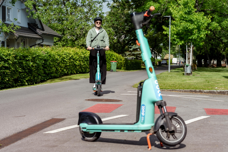 Im Vordergrund steht ein türkiser Leih-E-Scooter. Im Hintergrund fährt eine Frau mit Helm und Sonnenbrille auf einem E-Scooter. 