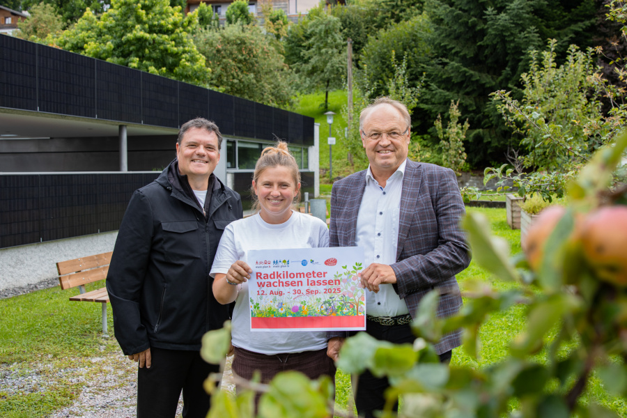 Bgm. Thomas Schierle, Julie Buschbaum und Bgm. Elmar Rhomberg stehen im Grünen und laden gemeinsam mit dem Plakat "Radkilometer wachsen lassen von 12. August - 30. September" zur Aktion. Im Vordergrund sind unscharf einige Äpfel am Baum zu sehen. Die Stimmung ist freundlich und naturverbunden.