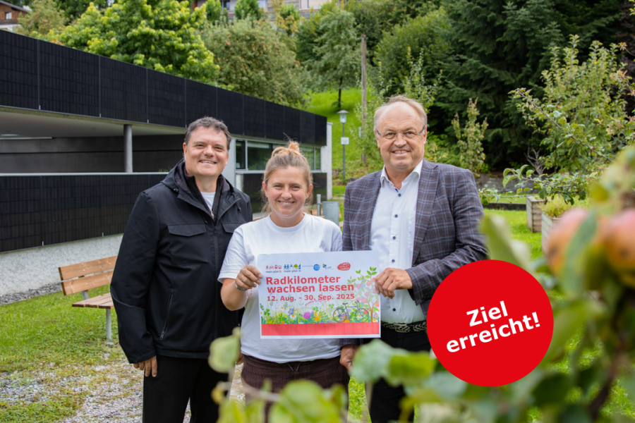 Bgm. Thomas Schierle, Julie Buschbaum und Bgm. Elmar Rhomberg stehen im Grünen und laden gemeinsam mit dem Plakat "Radkilometer wachsen lassen von 12. August - 30. September" zur Aktion. Im Vordergrund sind unscharf einige Äpfel am Baum zu sehen. Die Stimmung ist freundlich und naturverbunden.