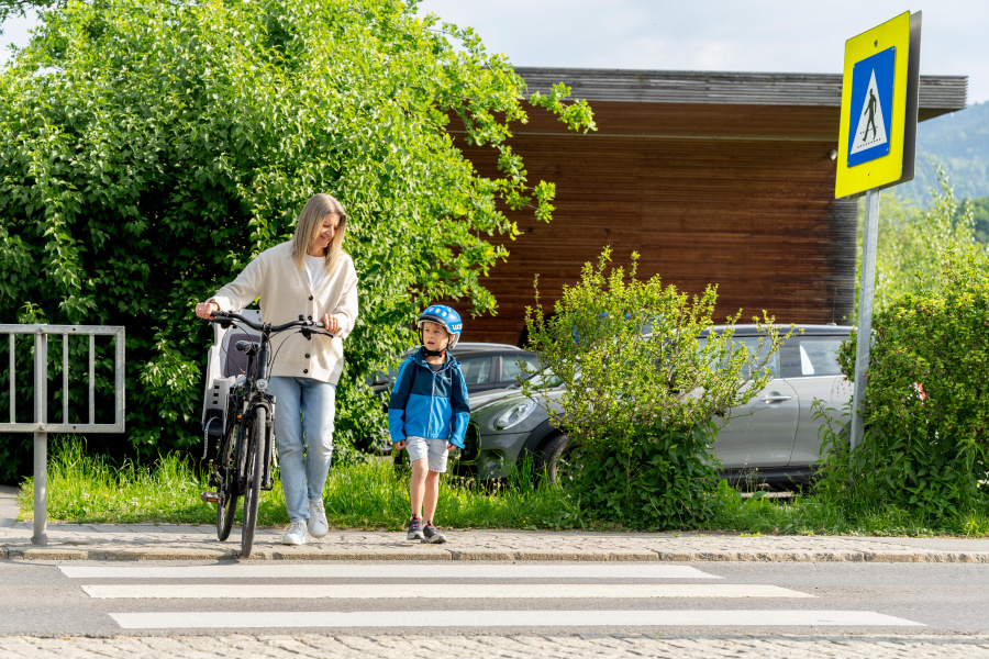 Eine Frau schiebt ihr Fahrrad über einen Zebrastreifen und begleitet dabei einen kleinen Jungen mit Fahrradhelm. Beide stehen am Straßenrand und wollen gemeinsam die Straße überqueren. Im Hintergrund sind grüne Sträucher, ein silbernes Auto und ein Holzgebäude zu sehen. Ein Verkehrsschild markiert die Fußgängerüberquerung