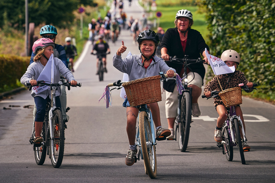 Eine Gruppe von Menschen fährt auf einer Landstraße mit Fahrrädern. Im Vordergrund fahren mehrere Kinder mit Helmen und Fahrradkörben, eines davon zeigt lächelnd den Daumen nach oben. Hinter ihnen folgen weitere Radfahrerinnen und Radfahrer. Es ist ein sonniger Tag und die Stimmung wirkt fröhlich und gemeinschaftlich.