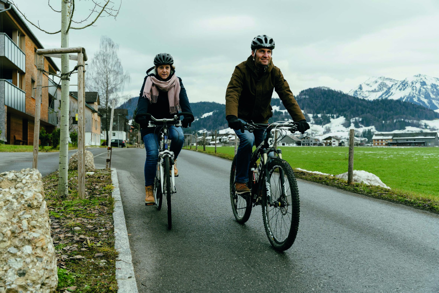 Zwei Personen sind warm gekleidet im Winter mit dem Fahrrad unterwegs. Im Hintergrund verschneite Berge. Die Straßen sind frei von Schnee. 